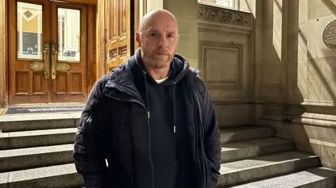 Striking bin worker Matthew Reid outside Birmingham Council House. He has a shaved head and a short blond beard. He wears a dark puffy jacket over a dark hoodie. The grey stairs behind him lead to wooden double doors.