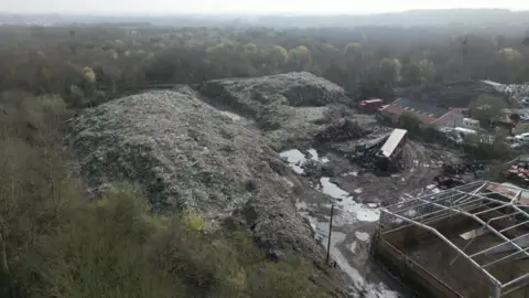 Two large mounds of rubbish, a building, a lorry and a derelict building can be seen at the site.