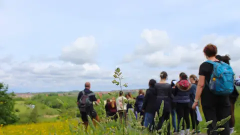 A group of young people walking at Rainton Meadows Nature Reserve. They are walking down a path which is surrounded by green space. Some have backpacks on their backs. The sky is blue with white clouds.