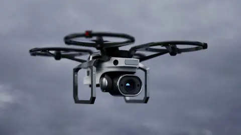 A grey and black drone hovering in the sky. It has four 'wings' as part of it. 