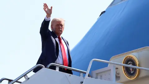 Reuters Donald Trump, wearing a dark suit, white shirt and red tie, waves as he emerges from the presidential plane Air Force One.