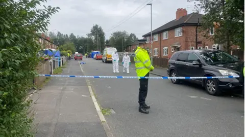 A police officer stands by police tape as forensics teams work at the scene.