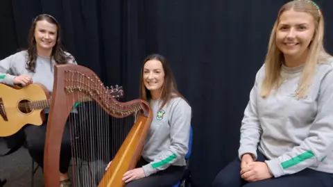 All-Ireland ballad group champions. Rachel Mitchell is posing with her guitar. One of her teammates has a harp and a third teammate is sitting with her hands in her lap