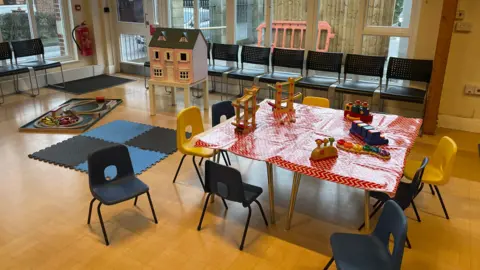 A table with blue and yellow chairs around it. There are a range of toddler toys on top of it. There is a pink dolls house in the background.