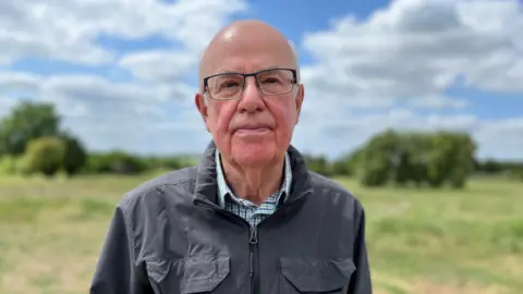 Robin Clegg looks into the camera wearing glasses and a blue jacket. He stands in front of the site, green with trees.