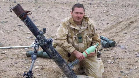 George Pas wearing beige and cream camouflage uniform and kneeling down in the dirt beside a shoulder fired missile launcher. 
