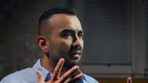 Olarte is wearing a blue shirt, his hands reaching out and related expressions while talking to the BBC