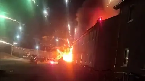 Fireworks go off from a burning van at the end of a row of terraced houses, the night sky is lit up by the fireworks