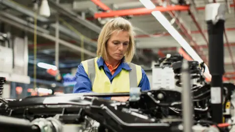 Getty Images A woman with short blonde hair wears high high-visibility vest as she looks down while working in a factory full of car parts