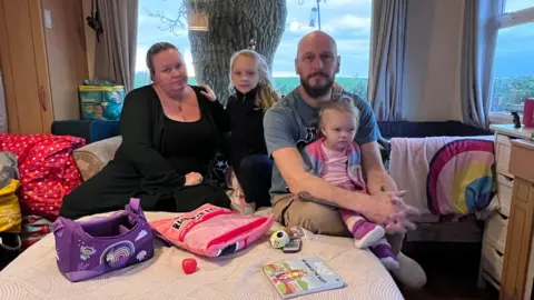 A family of four sat on a sofa bed in a caravan. There is a lot of stuff piled up in the corner to their left including a bag of nappies. A baby's cot is wedged to the right of the sofa.
On the bed the mum, dad and two daughters sit, one of the girls is a toddler.  There is some toys and books on the bed with them.