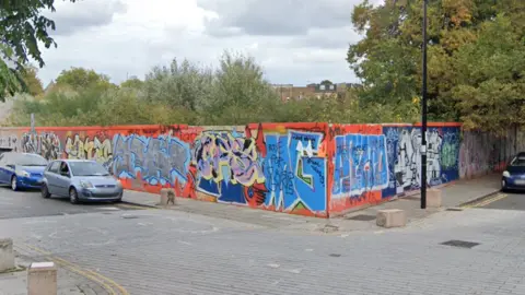 Google A street view image of orange hoarding around the waste ground site, which has graffiti sprayed on it. Cars can be seen parked on the roads around the site