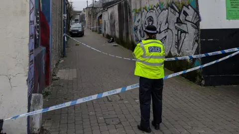 A police office wearing a yellow hi-vis jacket stood in front of blue and white police tape. The tape is covering an alley way with graffiti on the walls. 