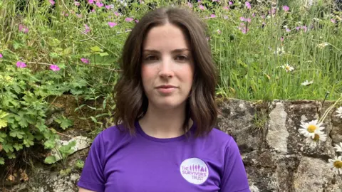 Leanne Rinne/BBC A woman with dark, short hair wearing a purple running T-shirt with a logo that reads 'The Survivors Trust'