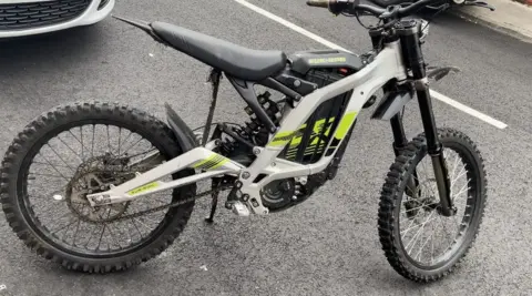 An electric bike on its stand in a car park. It has a white and yellow livery and the words "SUR RON" printed near the seat. The wheels have a thick tread. 