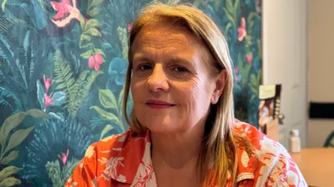 BBC Samantha Panton smiling for the camera. She is in her cafe. There is flowery wallpaper behind her. We can see a menu and some salt and pepper shakers blurred out in the background. She has long blonde hair and is wearing a colourful pink and white top. 