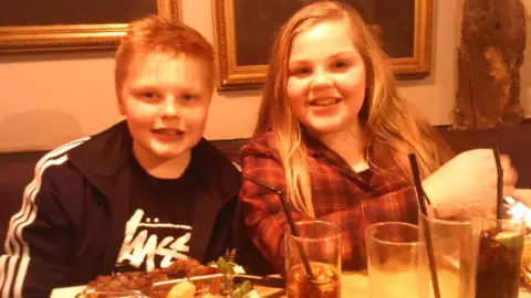 Handout A young Katie wears a red checked top and has long brown hair, her brother Harrison wears a black jacket and Stussy t-shirt. The pair are sat at the table, which has food and drinks on it, and smile at the camera