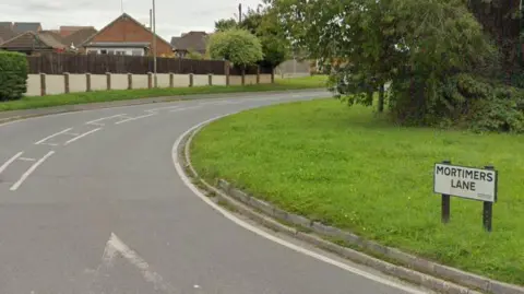 Mortimers Lane turning. The road sign is on a grass bank to the right of the road.