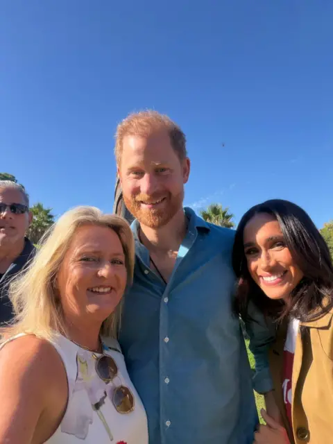 BBC/ Simon Atkinson A woman with blonde hair in a white top smiling with Harry and Meghan.