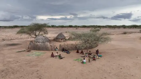 A group of people sit on the sandy ground outside a fenced enclosure with two thatched huts . The land looks very dry with just a few trees dotted around.