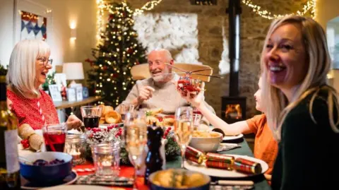 A family sit around the dinner table at Christmas. They are smiling and laughing. The child is holding a toy air ambulance.