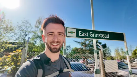 A man, Matt Nelson, stood next to a train station sign which reads "East Grinstead".