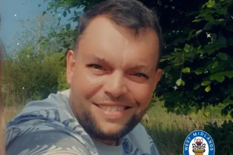 West Midlands Police A man with short brown hair and a short beard is sitting by trees in a field and smiling. He is wearing a blue patterned t-shirt.