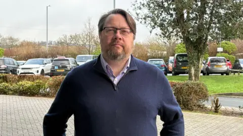 Dan has dark hair and glasses and a beard and wears a blue shirt and dark blue quarter-zip jumper. There are cars parked behind him and a hedge and trees. 