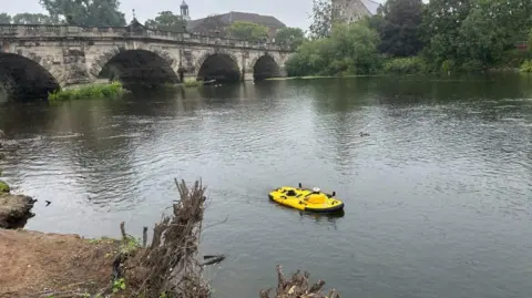 BBC/Charlotte Benton A bright yellow paddle board on a river near to the bank. A tree root is protruding from the bank, in line with the board. A stone bridge is behind with large archways cut out. 