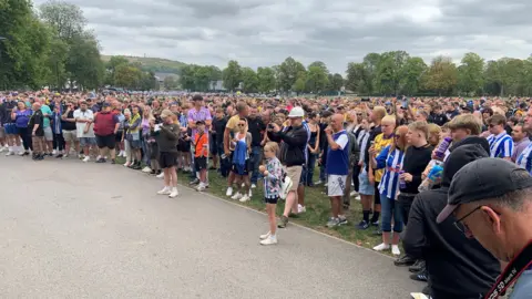 Hundreds of football fans, some in SWFC shirts, are gathered in a park - they form a large crowd and some are taking photos of the protest. 