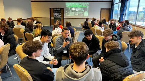 Dave Harvey/BBC Dozens of students attending a careers fair at Cheltenham Racecourse sit in groups with their chairs facing each other, chatting. They are in a large room with plain beige-coloured walls and blue carpet