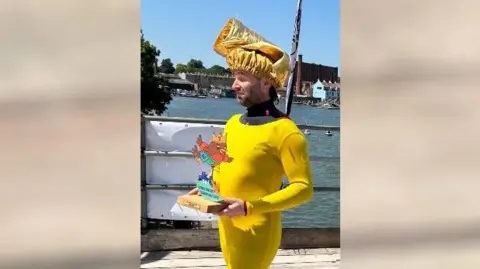 A man in skin-tight yellow clothing with a golden chef's hat on his head. He is carrying a winner's trophy