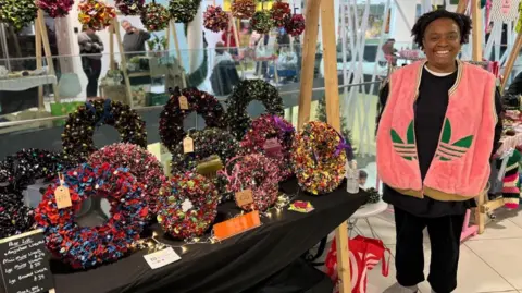 BBC A woman dressed in black and a pink gilet with green Adidas logo standing next to a stall of floral and foliage Christmas garlands and wreaths. There are also multi-coloured floral pom poms hanging form the stall. Other traders can been seen by stalls in the building behind her.