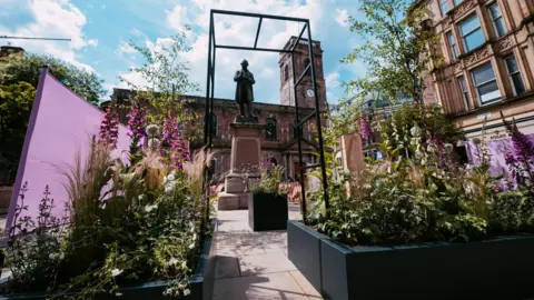 Plants and flowers line both sides of an arch over a narrow path in St Ann's Square. The old two-storey church with its clock tower is in the background with a statue in front seen between the arch.