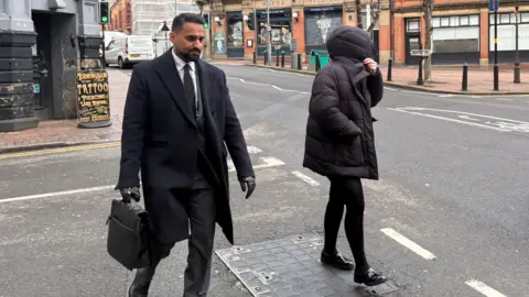 A man in a suit walks down a street carrying a briefcase, next to a woman who shields her face from the camera with the hood of her black puffer coat.
