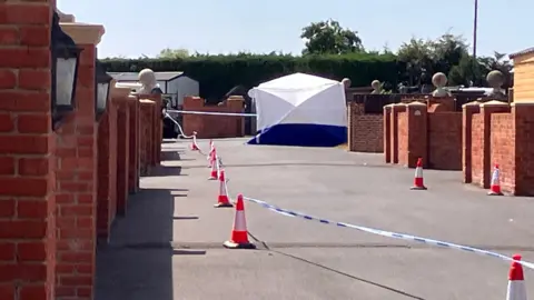 A white and navy forensics tent is set up on a travellers' site, with red brick walls either side of the shot. There are cones on the ground with police tape attaching them to each other.