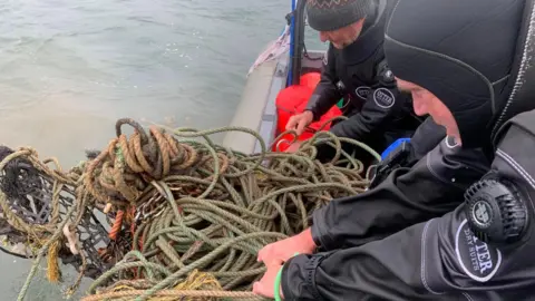 Clean Planet UK A huge tangle of rope and netting is being pulled out of the sea by two divers in waterproof gear. The rope is a green colour with a black net attached which is just emerging from the water.
