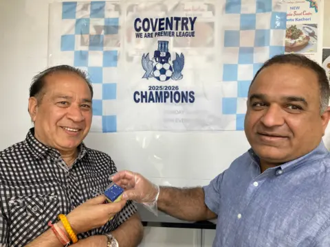 Surinder is standing on the left and is wearing a brown and cream checked shirt, on the right of him is jethaji odedra and he is wearing a chambray blue shirt and is handing a sky blue barfi to surinder. Between the two men is a blue and white flag with the words coventry champions.