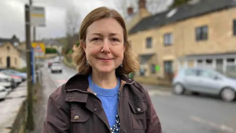 A middle aged woman with a brown bob smiles slightly as she stands on a damp street in a Cotswold village. There are cars driving behind her and a bus stop. She is wearing a brown coat with a blue jumper beneath it and a large silver necklace.