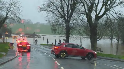 A line of traffic stands on a road which has been flooded. A large patch of water is visible in the distance with people trying to wade through and several cars driving through it. The road is flanked by several trees.