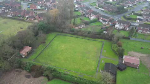 A drone view of a square bowling green with short green grass, surrounded by trees and several wooden and brick cabins. Houses can be seen a the top of the picture.