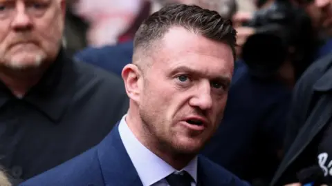 Reuters Image shows Stephen Yaxley-Lennon, known as Tommy Robinson, wearing a navvy blue suit, shirt and tie stands outdoors speaking while surrounded by a crowd of people which is slightly out of focus