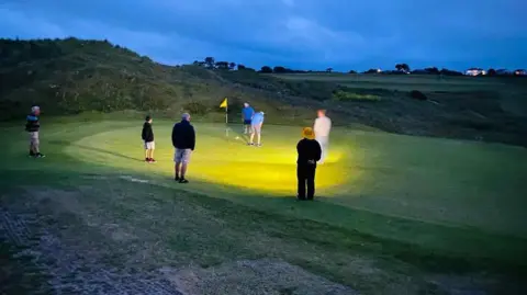 Two men play golf in the dark, using floodlights 