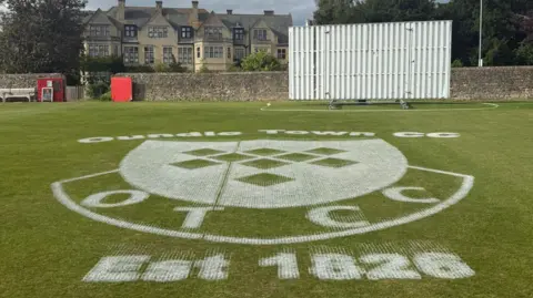Andrew Radd/BBC The logo of Oundle Town Cricket Club painted in white onto a cricket pitch. In the background a sight screen is visible at the boundary of the pitch.