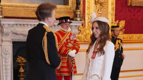 A side shot of Princess Anne, wearing a black uniform, talking to a young woman, who is wearing a white trouser suit. In the background are two men in uniform, including a Beefeater in red.