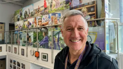 Andrew Turner/BBC Stuart Allen has medium length grey hair, slightly receding and swept over to his right. He is smiling, and is wearing a black hoodie and a purple lanyard around his neck. He is standing near a photomontage of children participating in school activities, set behind a glass ballustrade.