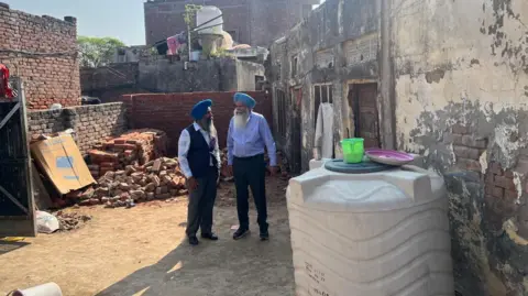 Paul Uppal Two men in turbans and suits stand outside a dilapidated building with peeling paint and piles of bricks outside.
