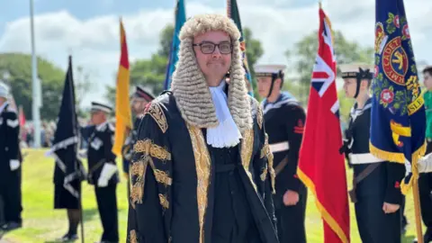 Speaker of the House of Keys Juan Watterson, who is wearing black and gold robes and a long white wig. There are standard bearers lined up alongside the walkway behind him.