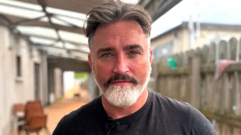 Brian Kerr has greying brown hair pushed back from his forehead. He has a neatly trimmed beard which is grey, but the mustache part of it is still mostly brown. He is standing in front of a wooden fence and there is a porch above his head which has clear perspex panels.