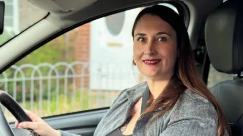 Elousie Kinsey sitting at the wheel of a car. She has long brown hair and is wearing a grey top and a light grey cardigan.