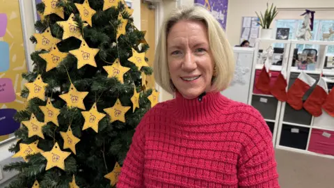 Tiggy McCool, standing in front of a large Christmas tree. She has short blonde hair and is wearing a pink turtleneck jumper. The tree is decorated with yellow stars. 